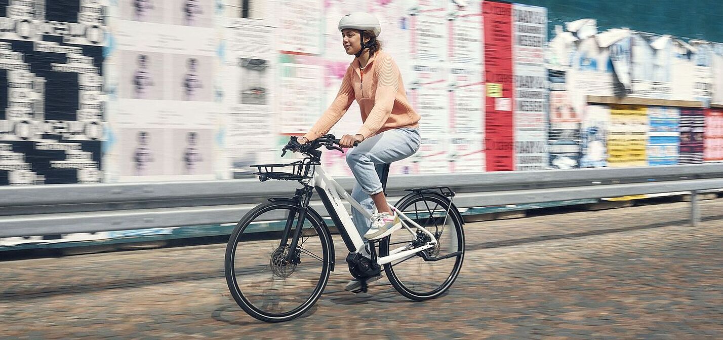 Elektrorad gebraucht Eine Frau fähet auf einem weißen Elektrorad vor einer Wand mit vielen Plakaten.