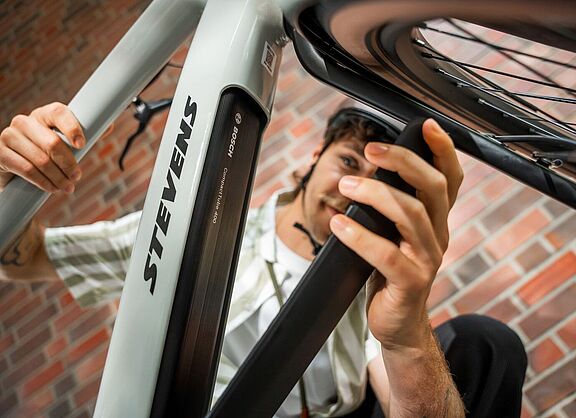 Akku Ein Mann öffnet die Akku-Abdeckung an einem grauen Elektrorad, von schräg unten aufgenommen.