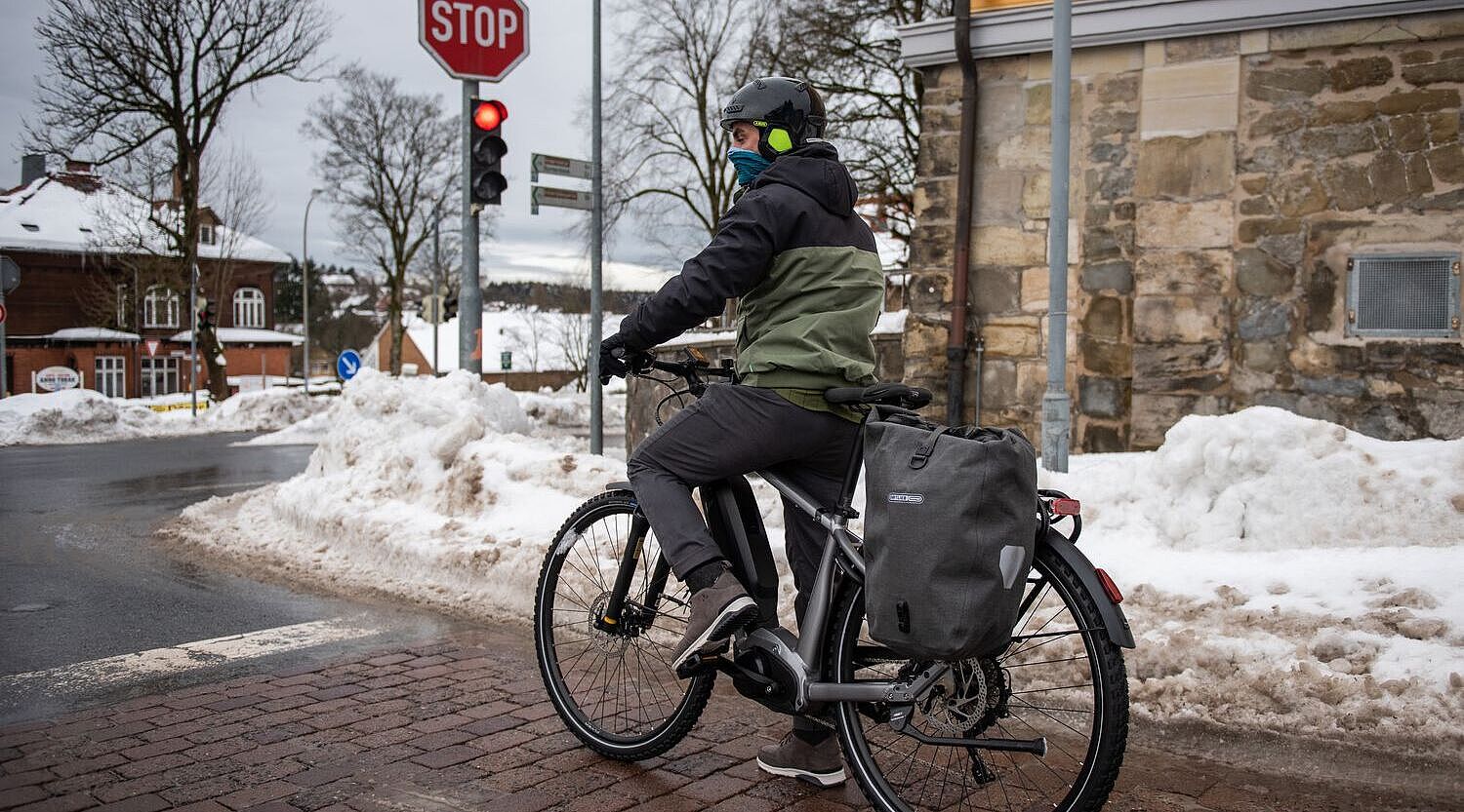Ein Mann steht mit Fahrrad an einer Ampel in winterlicher Stadtlandschaft. 